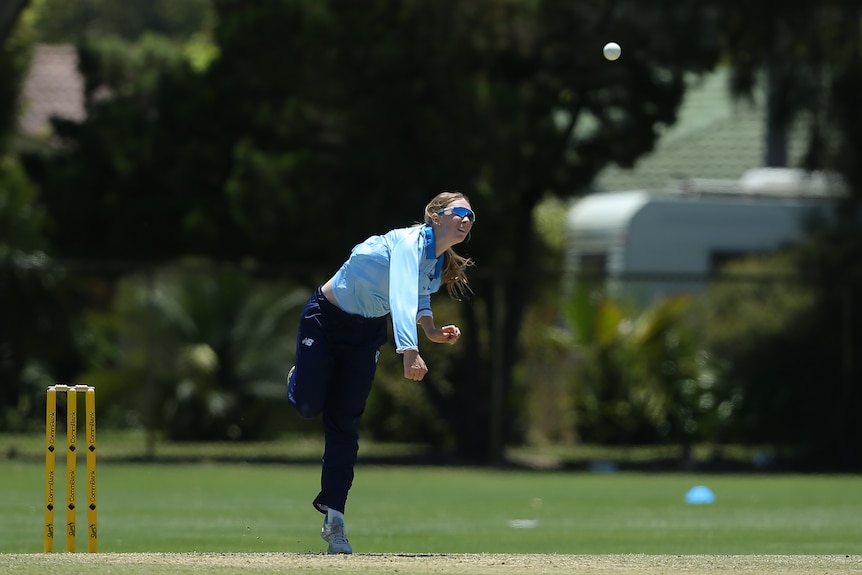 Women wearing long sleeved blue shirt bowls a ball in front of yellow cricket stumps