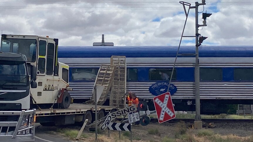 A truck with a damaged rear lies next to a train carriage with the word "Overland" on it.