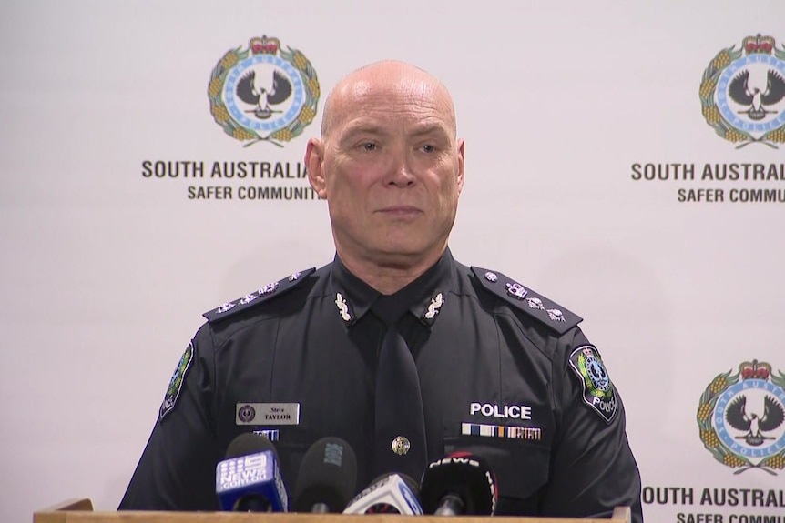 A man in police uniform speaking into microphones in front of sa police logo