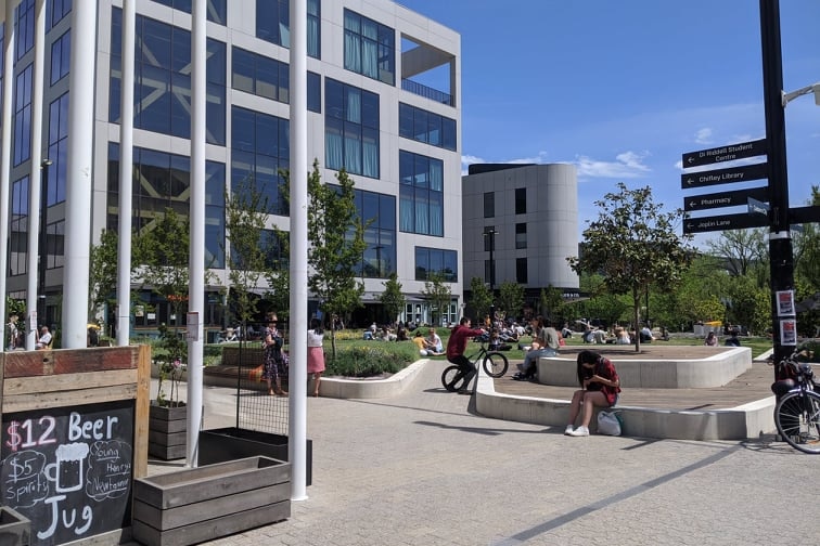 People sit and eat or walk, while a person cycles, through the campus of ANU. A sign says "$12 beer".