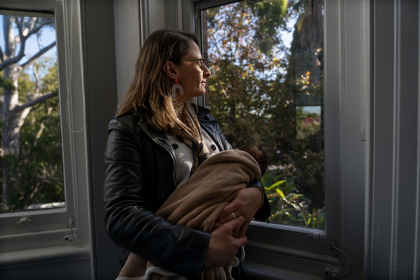 A woman holds a baby and looks out the window.