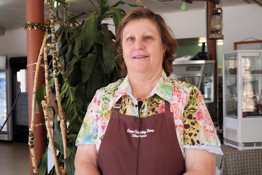 A middle aged woman with curly, shoulder length, auburn hair, wearing an apron, smiles at the camera.