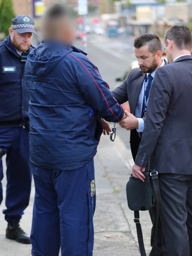 A detective places handcuffs on a man as another detective and uniformed policeman look on.