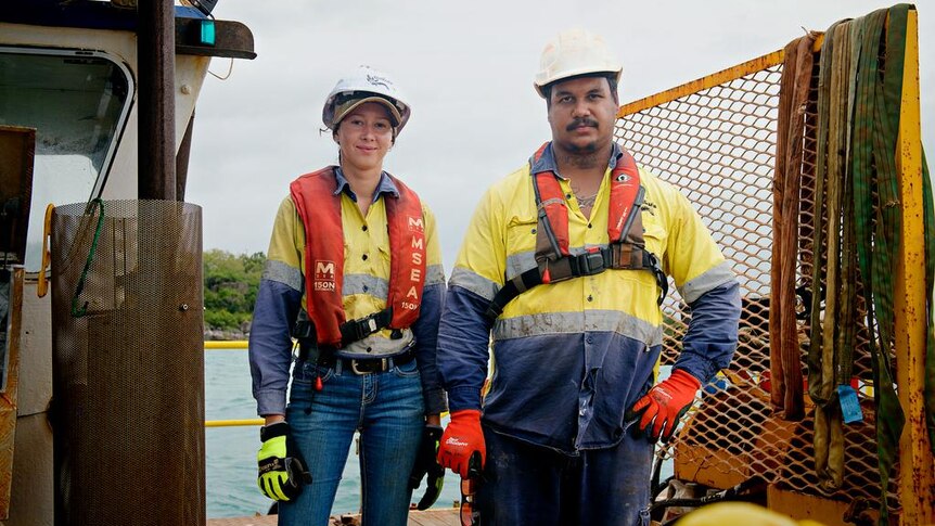 Two young people standing on a fishing-type boat, wearing high-vis shirts and life jackets. They are both smiling, slightly.