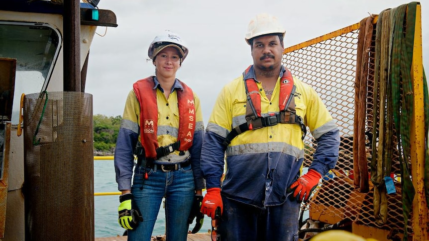 Two young people standing on a fishing-type boat, wearing high-vis shirts and life jackets. They are both smiling, slightly.