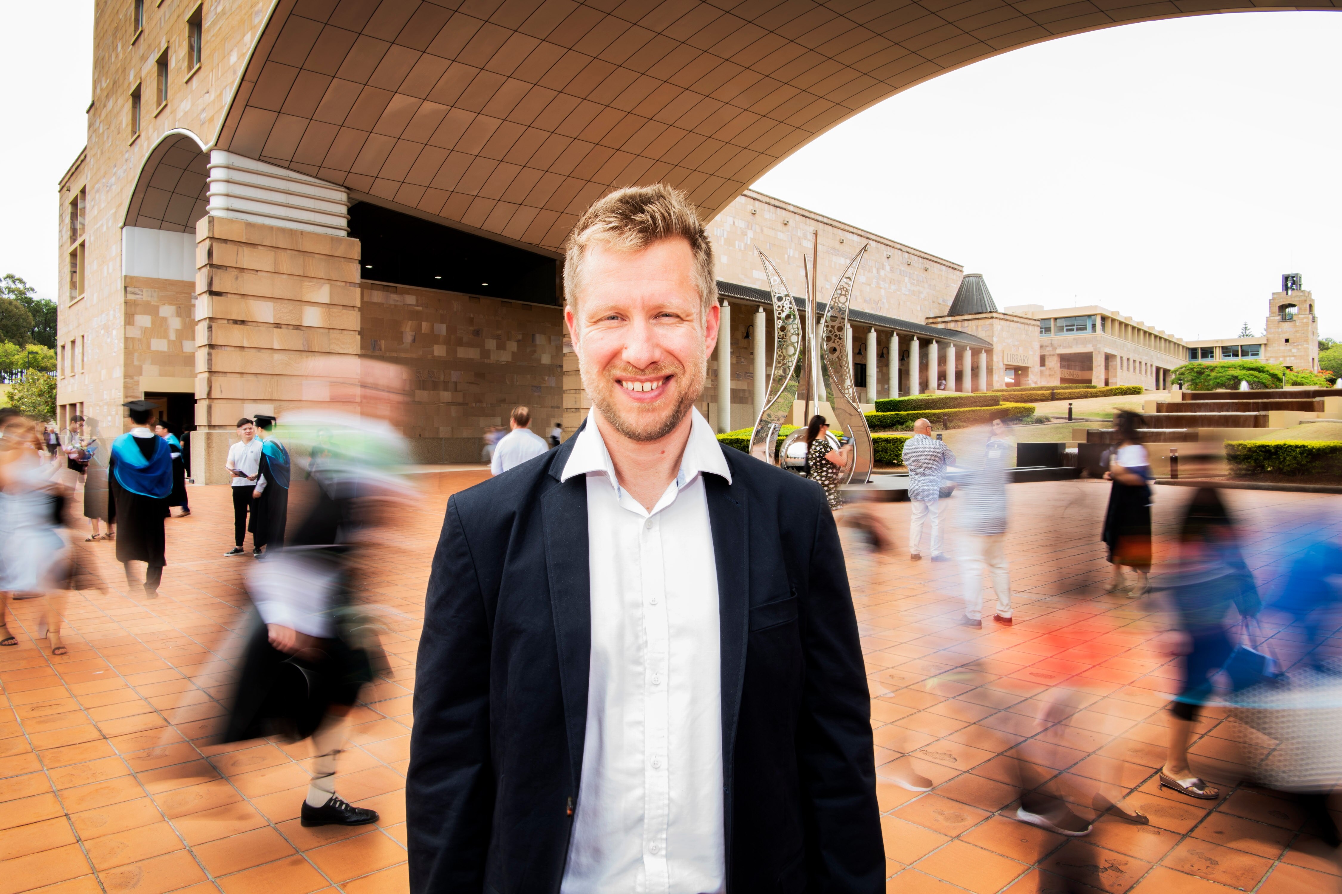 A man stands between sandstone buildings with students walking by