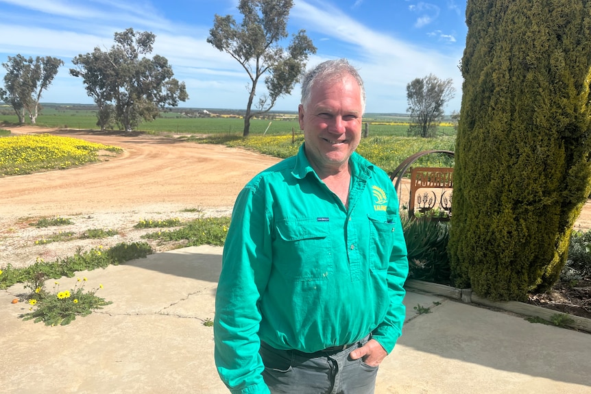 A man, wearing a green shirt, stands outside his house and smiles.