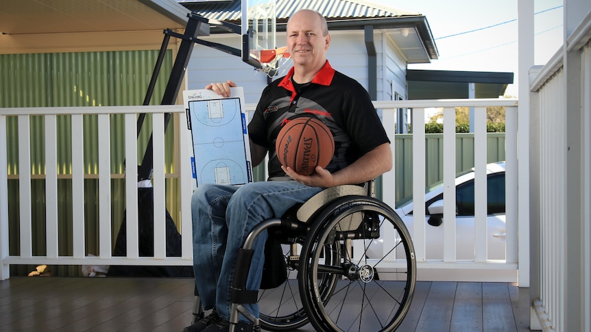 A smiling man in a branded polo shirt sits in a wheelchair holding up a small whiteboard with a basketball court image on it.