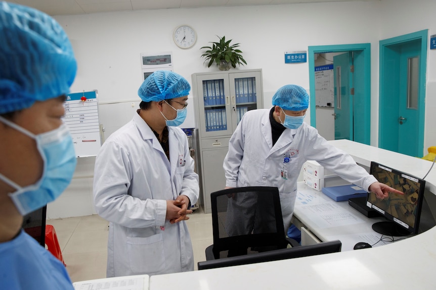 Three doctors wearing lab coats and personal protective equipment pointing at a screen