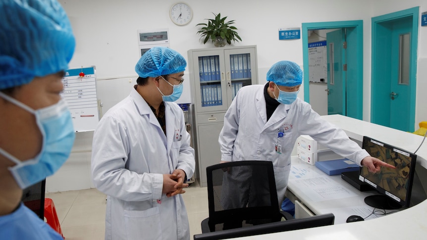 Three doctors wearing lab coats and personal protective equipment pointing at a screen.