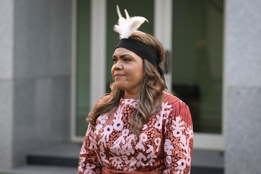 NT Senator Jacinta Price, wearing a feather headdress, standing outside a building and looking serious.