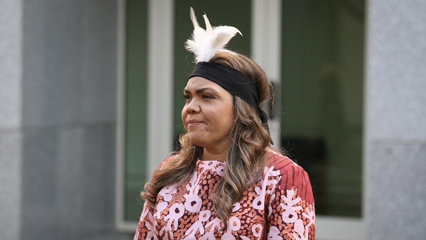NT Senator Jacinta Price, wearing a feather headdress, standing outside a building and looking serious.