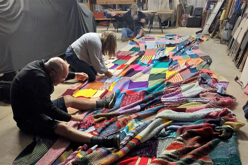 People hunched over a huge length of coloured knitted squares in a garage
