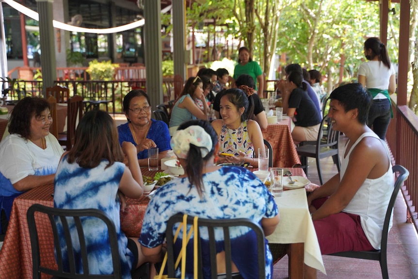 A group of women and men sit at tables in a restaurant surrounded by trees as waitresses walk nearby