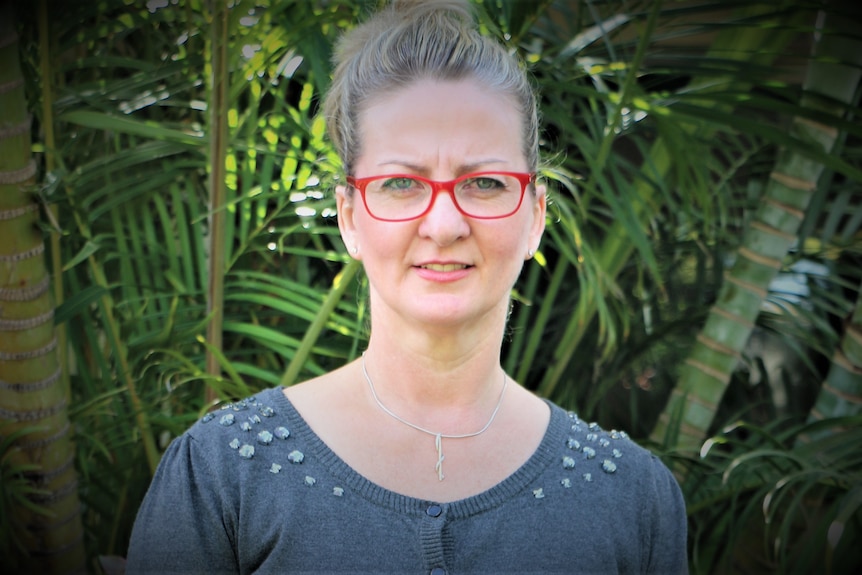 A blonde woman with red glasses looks at the camera from in front of a leafy background.