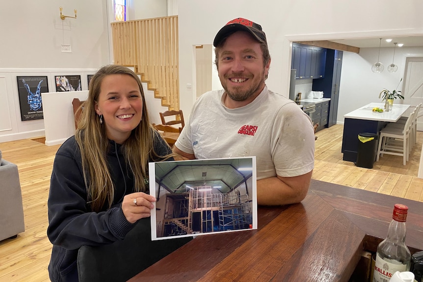 A woman and a man stand leaning on a bar holding a photo of a construction site. 