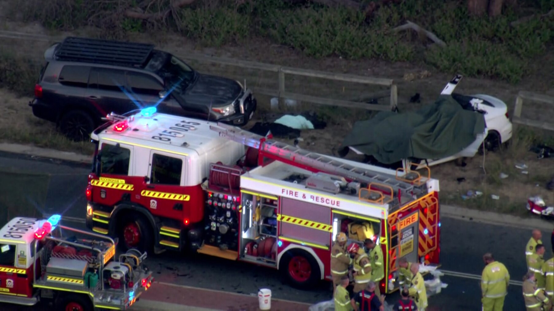 An aerial shot showing a crowd of firefighters and a fire truck next to the wreckage of a white car after a road crash.