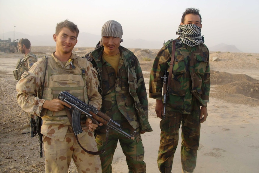 Three man smile as they stand in a sandy barren landscape dressed in military camouflage fatigues and holding rifles