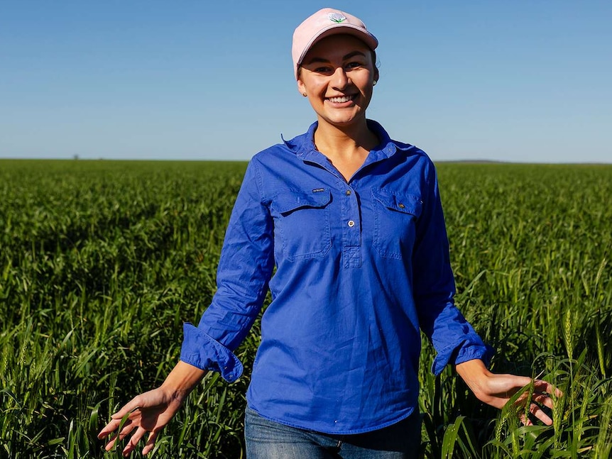 A young woman walks through a sorghum crop, running her hands through it as she goes. Blue sky in background.