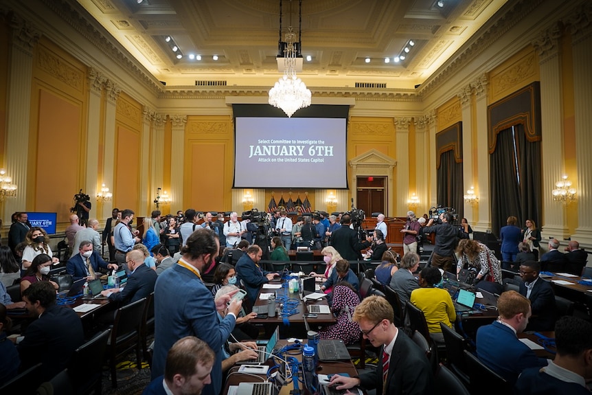 A crowded room with a big chandelier and grand white columns, with "January 6th" written on a screen in the centre