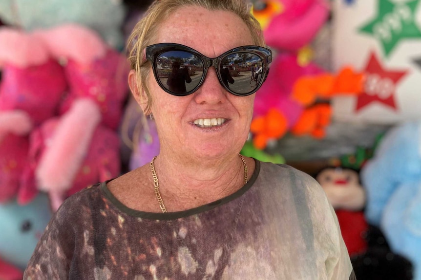 A blonde woman wearing large black sunglasses stands in front of a carnival attraction.