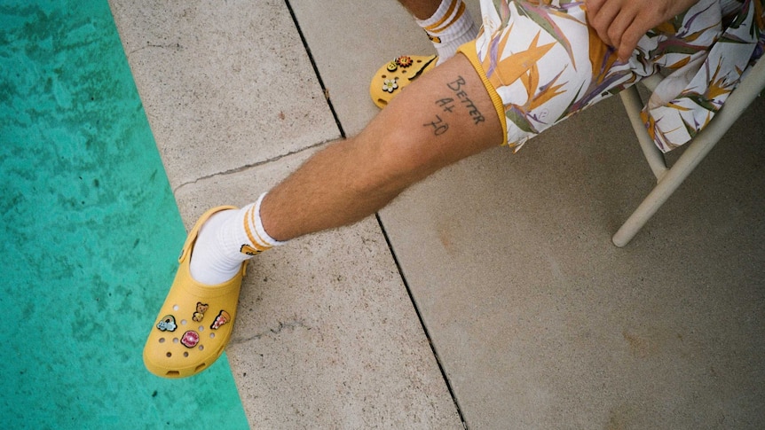 A leg, in shorts, next to a pool, with socks and yellow plastic Crocs on the feet. The leg has a tattoo that says "better at 70"