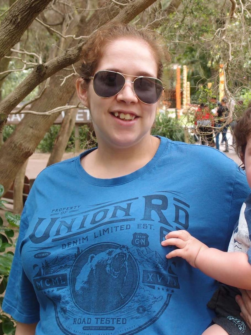 A woman wearing sunglasses with a toddler on her hip smiles at the camera.