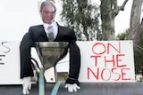 An effigy in a black suit with a picture of mans face sits in toilet next to protest signs