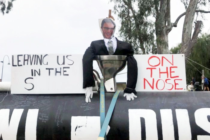 An effigy in a black suit with a picture of mans face sits in toilet next to protest signs