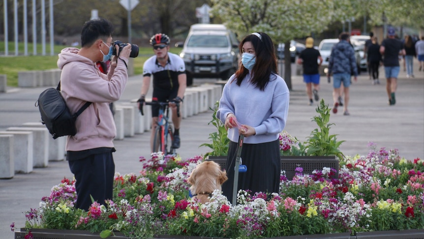 A man takes a photo of a woman in front of flowers.