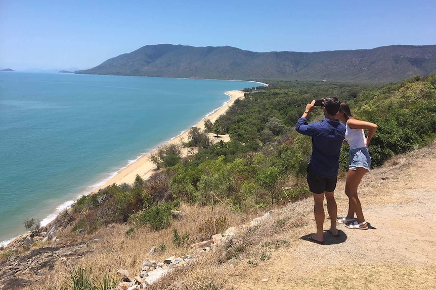 A man and a woman use a mobile to take a photo  of the scenic view of a beach and greenery.