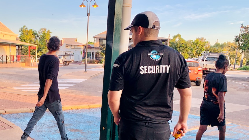 A male security guard stands with his back to the camera as shoppers walk past.