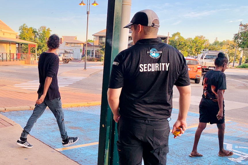 A male security guard stands with his back to the camera as shoppers walk past.