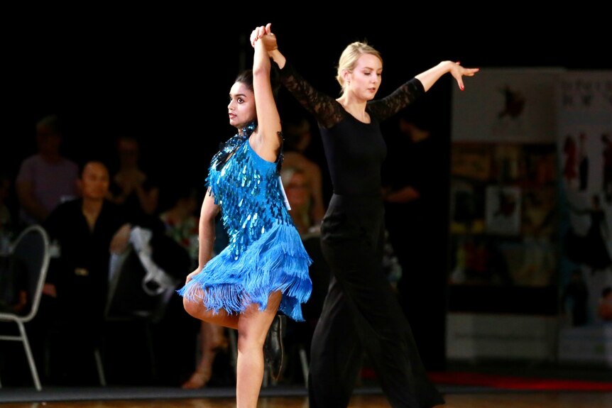 A girl wearing a blue dress takes part in ballroom dancing with the aid of a woman dressed in black.