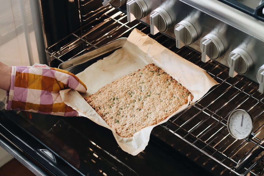 A gloved hand pulls a muesli slice, on a paper-lined tray, out of an oven.