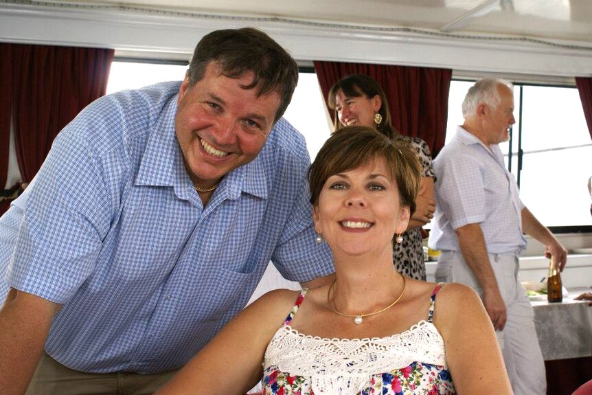 A man in a blue checked shirt leans in towards his seated wife, both smiling at the camera.