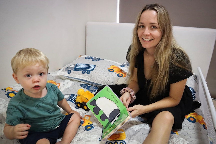 A woman sits with her son on a bed reading a book.