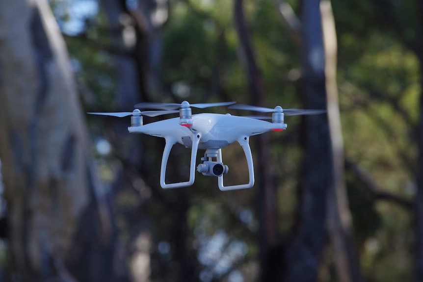 A flat white drone with four rotors in flight with trees in the background.