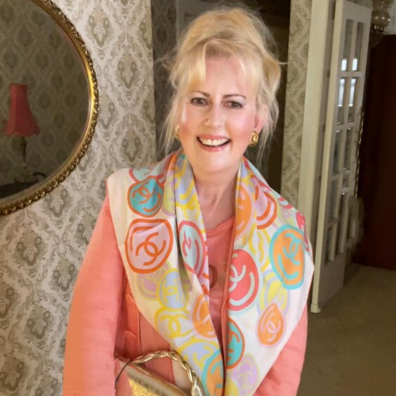 A blonde woman is wearing pink with a patterned scarf and gold handbag. 