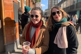 Two women wearing scarves and jackets and sunglasses smile as they hold a glass of beer in their hands