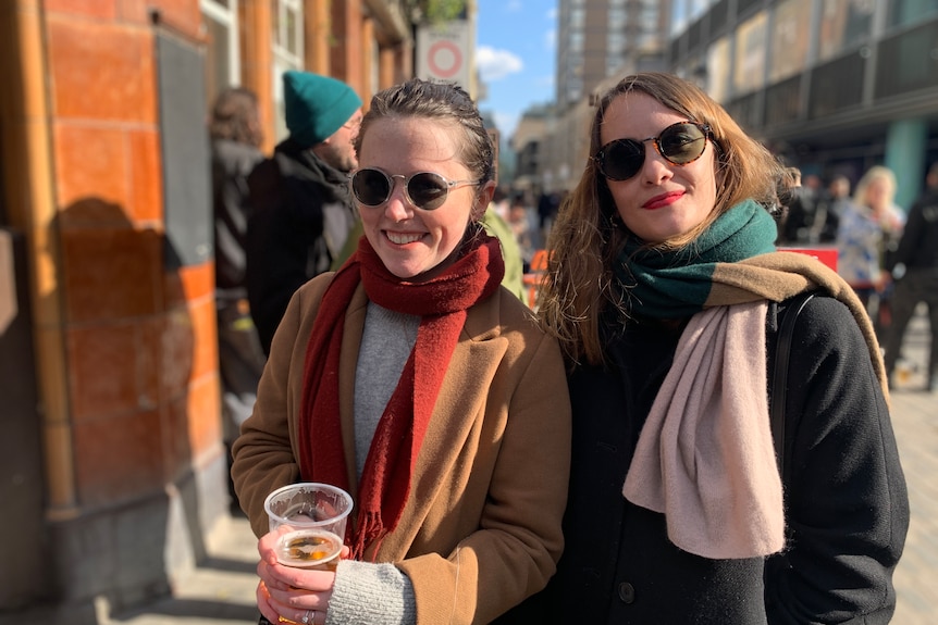 Two women wearing scarves and jackets and sunglasses smile as they hold a glass of beer in their hands