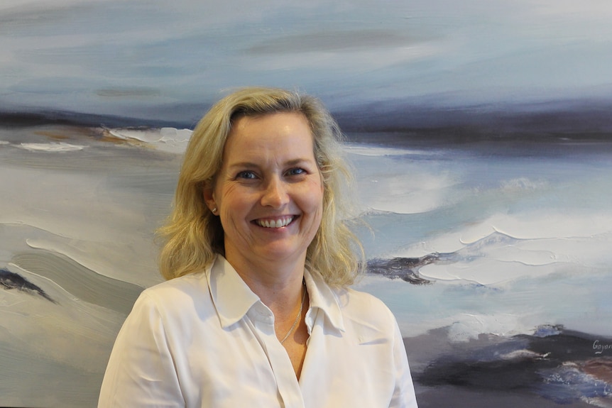A blonde haired woman smiles at the camera in a white shirt in front of an ocean painting