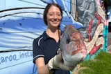 Woman holds a whole fresh fish up toward the camera. 