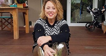 A woman, sitting on an outdoor deck with table and pram in the background, holds champagne glass and smiles broadly.