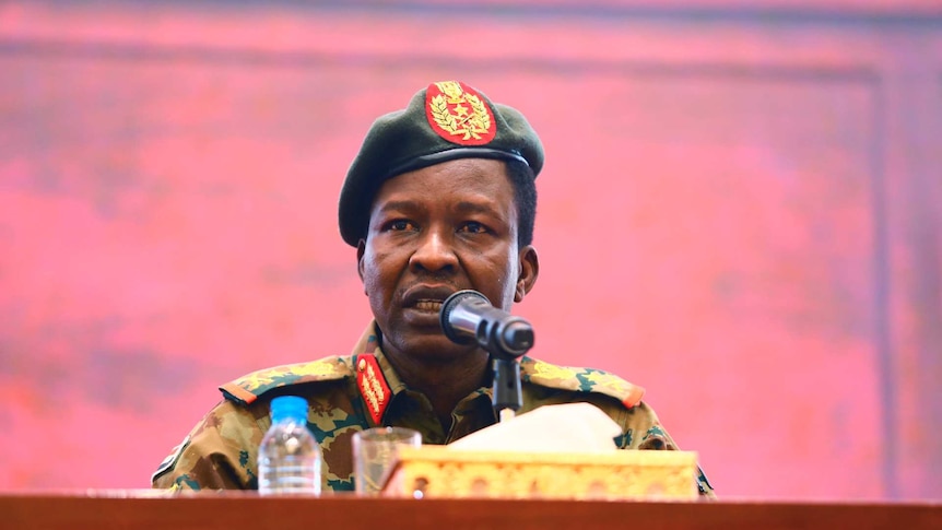 Sudan's ruling Military Council spokesperson speaks in front of a microphone in army uniform in front of a pale pink background.