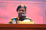 Sudan's ruling Military Council spokesperson speaks in front of a microphone in army uniform in front of a pale pink background.