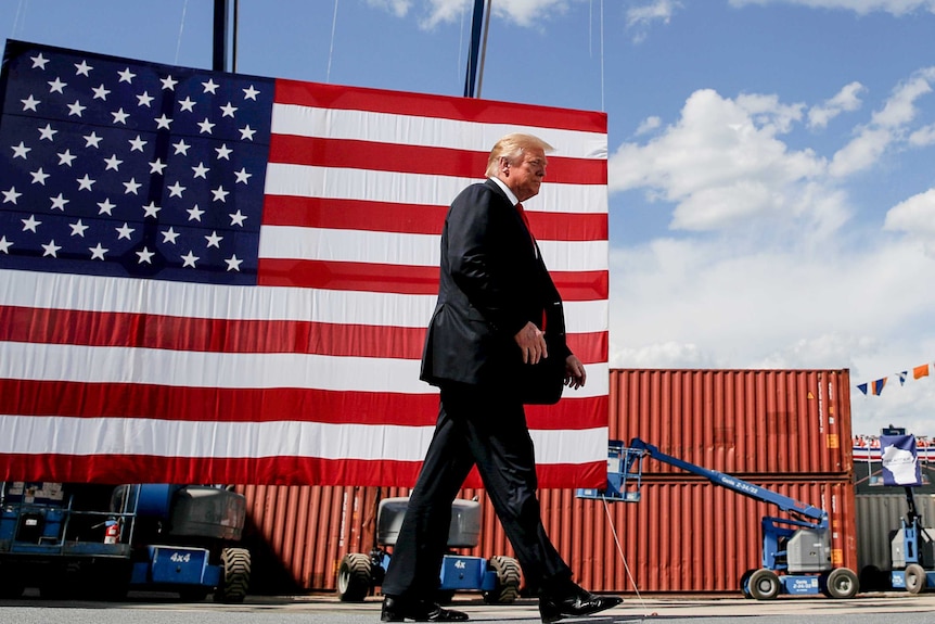 Donald Trump in a suit and red tie walking past an American flag