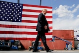 Donald Trump in a suit and red tie walking past an American flag