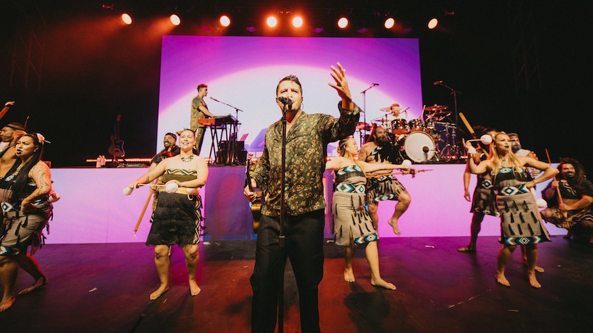 A man sings on a stage raising one hand with maori kapa haka performers in traditional dress dancing in the background 