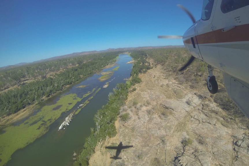 An aerial view of a river, with a light plane with a propeller in the sky to the right, and its shadow below.